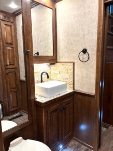 Small bathroom with wood cabinets, a vessel sink on a stone countertop, a black faucet, tiled backsplash, large mirror, towel ring, and beige textured walls. Part of a toilet and wood flooring are also visible. -Stillwater Trailer Sales