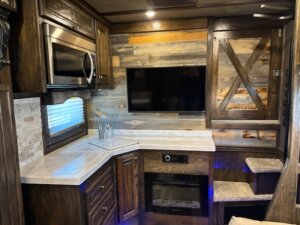 Cozy RV kitchen with dark wood cabinets, marble countertops, a mounted microwave, sink, TV, electric fireplace, and stairs with carpeted steps leading up to another area. Rustic wood paneling decorates the walls. -Stillwater Trailer Sales