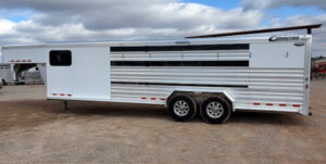 A large, silver and white Cimarron stock combo livestock trailer with dual axles is parked on gravel under a cloudy sky. This 26' trailer features horizontal metal paneling and a convenient side entry door. -Stillwater Trailer Sales
