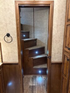 A wooden door opens to reveal carpeted stairs with small blue LED lights on each step, leading upward in a cozy, wood-paneled interior space. -Stillwater Trailer Sales