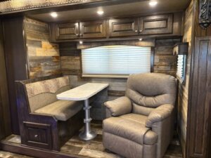 A cozy RV interior features a padded dining booth with a marble-style table and overhead cabinets, wood-paneled walls, blinds on the window, and a large, plush recliner chair next to the booth. -Stillwater Trailer Sales