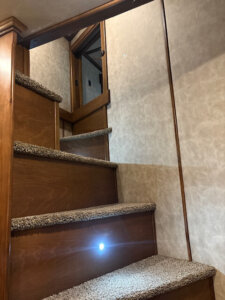A carpeted staircase with wooden sides and a small built-in light leads up to a closed wooden door in a beige-walled interior space. -Stillwater Trailer Sales