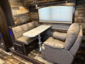 A cozy RV dining area with a cushioned booth on one side of a rectangular table and a beige reclining chair on the other, set against rustic wood-paneled walls and a window with closed blinds. -Stillwater Trailer Sales
