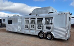 A large silver gooseneck livestock trailer with five barred windows and vents, dual wheels, and roof-mounted air conditioning units parked on a dirt lot under a partly cloudy sky. -Stillwater Trailer Sales