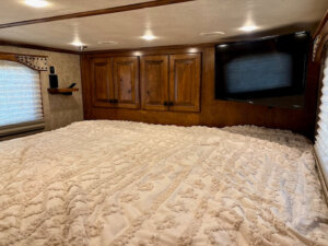 A cozy bedroom with a large bed covered in a textured white blanket, wooden cabinets on the wall, a flat-screen TV, and windows with blinds letting in soft natural light. -Stillwater Trailer Sales