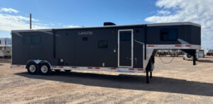 A large, dark gray Lakota gooseneck trailer with white trim is parked on a gravel lot under a partly cloudy sky. It has multiple windows, dual axles, and an elevated front section. -Stillwater Trailer Sales