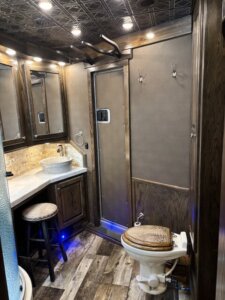 A rustic-style bathroom with dark wood cabinets, a vessel sink, a stool, a walk-in shower with a glass door, a toilet with a woven seat cover, and wood-patterned floor and ceiling panels. Warm lighting is overhead. -Stillwater Trailer Sales