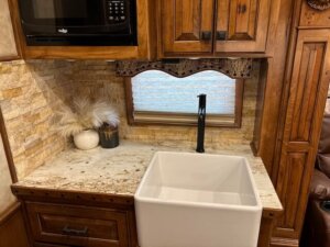 A kitchen countertop with a white farmhouse sink, a tall black faucet, brown wooden cabinets, a microwave, a window with a blind, and two decorative vases with dried grass next to the sink. -Stillwater Trailer Sales