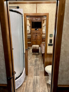 View from a doorway into an RV interior, showing a curved shower on the left, toilet on the right, wood floors, and a living area with a sofa, cabinets, and a TV in the background. -Stillwater Trailer Sales