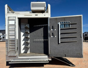 The image shows the open rear compartment of a 2019 Bloomer 5 Horse trailer, revealing saddle racks, bridle hooks, and storage bars on the inside of the door. The trailer is parked on a dirt lot under a clear blue sky. -Stillwater Trailer Sales