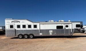 A 2019 Bloomer 5 Horse silver and white gooseneck trailer with six windows, triple axles, and rooftop AC units sits parked on a dirt lot under a clear blue sky. The trailer is branded BLOOMER TRAILERS. -Stillwater Trailer Sales