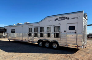 A large, silver 2019 Bloomer multi-horse trailer with six windows, triple axles, and &ldquo;Bloomer Trailers&rdquo; branding is parked on a gravel lot under a clear blue sky. Perfect for transporting up to 5 horses in style and comfort. -Stillwater Trailer Sales
