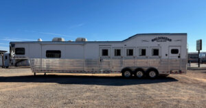 A large silver and white 2019 Bloomer gooseneck horse trailer with multiple windows and three axles is parked on a gravel lot under a clear blue sky. This spacious trailer is designed to accommodate up to 5 horses. -Stillwater Trailer Sales
