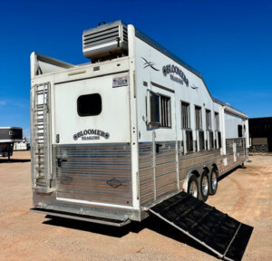 A large white 2019 Bloomer 5 Horse trailer with multiple windows and vents is parked on a dirt lot. The back door is open with a ramp extended to the ground. The sky is clear and blue. -Stillwater Trailer Sales