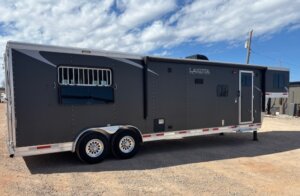 A large, black gooseneck horse trailer with silver trim is parked on a dirt lot under a partly cloudy sky. It has two axles, windows with bars, and a side door. -Stillwater Trailer Sales