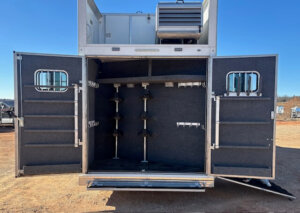 Open storage compartment of a horse trailer with carpeted interior, multiple vertical and horizontal racks for saddles and tack, and open double doors; ventilation unit visible on top under a clear blue sky. -Stillwater Trailer Sales