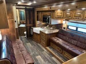 Interior of a rustic 2019 Bloomer horse trailer with wood paneling, two brown leather sofas, a kitchen area featuring a farmhouse sink and microwave, and a bathroom visible in the background. Natural light filters through window blinds. -Stillwater Trailer Sales