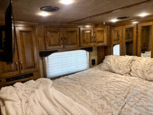 Cozy RV bedroom in a 2019 Bloomer 5 Horse with wood cabinets, a large bed covered in a cream textured blanket and pillows, a window with a shade, and a TV mounted on the wall. Warm lighting creates a comfortable atmosphere. -Stillwater Trailer Sales