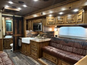 Interior of a cozy 2019 Bloomer RV with wood-paneled cabinets, a farmhouse sink, marble countertop, microwave, brown leather sofa, and sliding barn door to the bathroom. Natural light streams through a window with blinds in this 5 Horse model. -Stillwater Trailer Sales