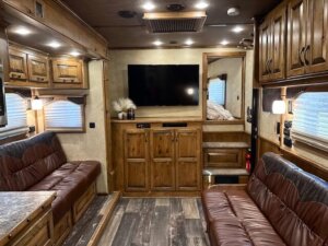 A cozy 2019 Bloomer RV interior with brown leather sofas, wood cabinets, a wall-mounted TV, and windows with blinds. The space features warm lighting, decorative items, and wood-patterned flooring&mdash;perfect for a 5 Horse setup. -Stillwater Trailer Sales