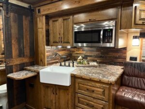 A rustic 2019 Bloomer kitchen features wooden cabinets, stone-patterned countertops, a farmhouse sink, microwave above, and a multi-toned wood backsplash. A sliding barn door opens to reveal part of the 5 Horse bathroom. -Stillwater Trailer Sales
