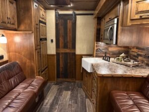 A cozy 2019 Bloomer RV interior with wood-paneled walls, leather seating, a small kitchen area featuring a farmhouse sink and marble countertop, plus a sliding barn door in the back. Designed for comfort after long 5 Horse adventures. Warm lighting adds to its welcoming feel. -Stillwater Trailer Sales