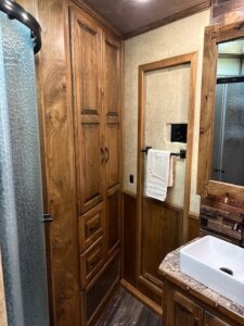 A compact Bloomer bathroom features wooden cabinets, a glass shower door, a sink with a marble-patterned countertop, a mirror, and a towel hanging on a rack attached to the wall. -Stillwater Trailer Sales