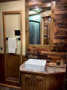 Rustic bathroom in a 2019 Bloomer 5 Horse trailer, featuring wood-paneled walls, a framed mirror, white vessel sink with black faucet, granite countertop, towel rack with white towel, and a shower visible in the background. -Stillwater Trailer Sales