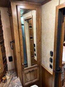 A wooden cabinet with a full-length mirror stands in the bathroom of this 2019 Bloomer 5 horse trailer, next to a granite countertop, hooks, light switches, and a small basket with toiletries. A shower with a glass door is visible in the background. -Stillwater Trailer Sales
