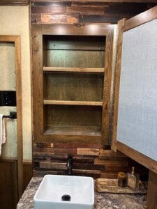 A wooden medicine cabinet by Bloomer with its door open reveals two shelves above a white sink and faucet. The countertop holds small decorative items, and 5 Horse wood panels from 2019 accent the wall. -Stillwater Trailer Sales