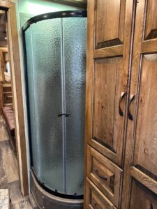 A compact bathroom in a 2019 Bloomer trailer features a corner shower with frosted glass doors and curved frame, next to tall wooden cabinets with dark handles, all set on wood-patterned flooring. -Stillwater Trailer Sales