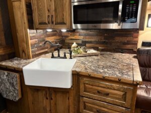 Rustic kitchen with a farmhouse sink, wooden cabinets, and a marble countertop&mdash;perfect for relaxing after a day on the road with your 2019 horse trailer. Wood plank backsplash and fresh herbs add cozy charm by the wall. -Stillwater Trailer Sales