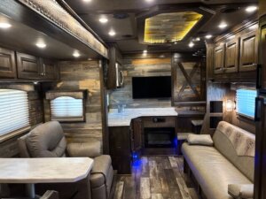 The image shows the interior of a modern RV with wood-paneled walls, leather seating, a small dining table, a wall-mounted TV, cabinets, a countertop, and stylish recessed lighting. -Stillwater Trailer Sales