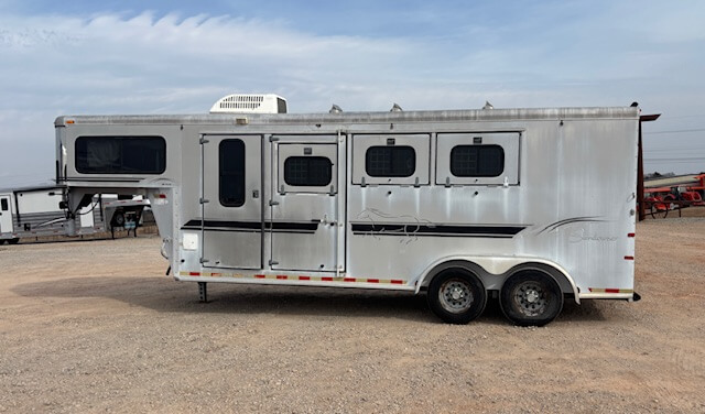 A silver Sundowner 3 Horse gooseneck trailer with three windows and a side door is parked on a gravel lot under a cloudy sky. The horse trailer features two axles and black trim details. -Stillwater Trailer Sales