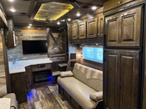 Interior of a modern RV with dark wood cabinets, a beige sofa, a small kitchenette, a wall-mounted TV, fireplace, and a window with a shade. The space has recessed lighting and rustic decor elements. -Stillwater Trailer Sales