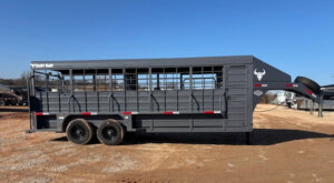 A large, dark gray Swift Built livestock trailer with a convenient Tack Box Combo, open bars along the sides, and tandem wheels is parked on a gravel lot under a clear blue sky. -Stillwater Trailer Sales