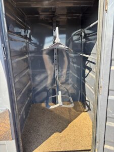 A small, dark Swift Built metal storage compartment with a textured floor and two vertical saddle racks mounted in the center. Light enters from the open door, casting shadows inside this sturdy Tack Box Combo. -Stillwater Trailer Sales