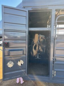 A blue Swift Built metal trailer with its side door open reveals an empty interior with saddle racks and a Tack Box Combo. Two small vents are on the door, and a pair of pink boots is visible peeking out from underneath. -Stillwater Trailer Sales