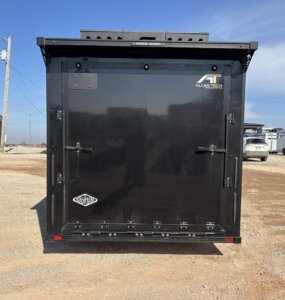 The image shows the back view of a large black bumper pull horse trailer parked on a gravel lot, with &ldquo;Alumitech&rdquo; and &ldquo;Outlaw&rdquo; logos on the door. Other vehicles and buildings appear in the background under a blue sky. -Stillwater Trailer Sales