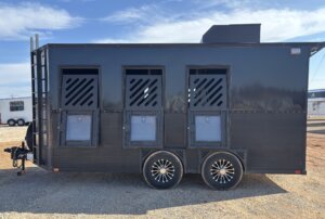 A black Alumi Tech horse trailer with three side doors featuring diagonal vents and transparent panels, two wheels per side, and parked on gravel under a blue sky. This 3 horse bumper pull offers both style and functionality. -Stillwater Trailer Sales