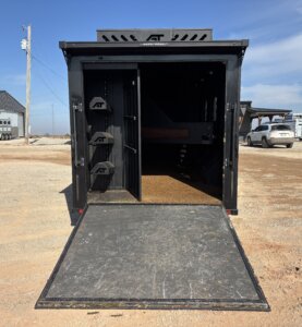 A black Alumi Tech 3 Horse Bumper Pull trailer with its rear door open as a ramp reveals an empty interior, saddle racks on the left wall, and a brown floor. The trailer is parked on gravel near other vehicles and buildings. -Stillwater Trailer Sales