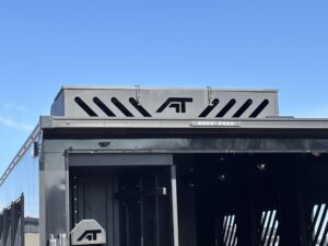 A close-up of the upper rear section of an Alumi Tech metallic black horse trailer, featuring the initials AT in a bold logo against a clear blue sky. The bumper pull trailer showcases modern, angular design elements and ventilation slits. -Stillwater Trailer Sales