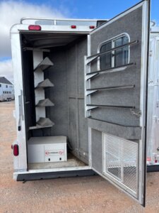 Open trailer door revealing a carpeted interior with four angled saddle racks on the left, a white Guardian Rx box on the floor, and ventilation panels on the door. The sky and part of another trailer are visible outside. -Stillwater Trailer Sales