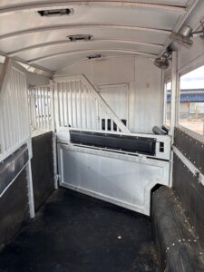 Interior of an empty aluminum horse trailer with a partition divider, padded side rails, rubber flooring, and a windowed side door leading outside. Natural light is coming in through the windows. -Stillwater Trailer Sales