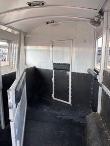 Interior of an empty horse trailer with metal walls, a black rubber floor, padded side panels, ventilation windows, and a closed rear door. The space is designed for transporting animals safely. -Stillwater Trailer Sales