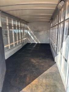 View of the interior of an empty 26' Cimarron Stock Combo livestock trailer with metal walls, ventilation openings on both sides, a curved roof, and a dark rubber floor. Sunlight casts shadows inside from the openings. -Stillwater Trailer Sales