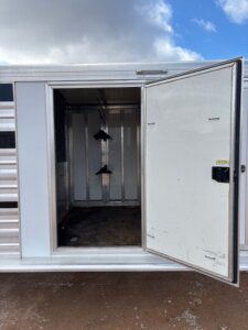 A white Cimarron Stock Combo trailer with an open side door reveals an empty interior featuring two black saddle racks on the wall. The ground outside is dirt, and the sky above is partly cloudy with patches of blue. -Stillwater Trailer Sales