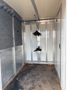 The image shows the empty interior of a 26' Cimarron Stock Combo horse trailer. Two black feed bins hang from the far wall, and bridle hooks line the left side. The metal floor is dirty and stained. -Stillwater Trailer Sales
