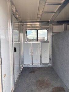 Interior of an empty 26' Cimarron stock combo horse trailer with a gray carpeted floor, white metal walls, a small window showing a partly cloudy sky, and a half-height divider on the right side. -Stillwater Trailer Sales