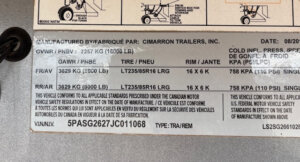 A metal label on a Cimarron stock combo trailer lists specifications such as manufacturer, date (08/2020), GVWR 16,000 lbs, axle ratings, tire sizes, PSI, VIN, and compliance details for this 26' model in English and French. -Stillwater Trailer Sales